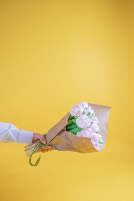 A person dressed in a white shirt is holding a bouquet of fresh light pink tulips with green stems and leaves, wrapped in light brown kraft paper and tied with a green ribbon. The bouquet is extended 