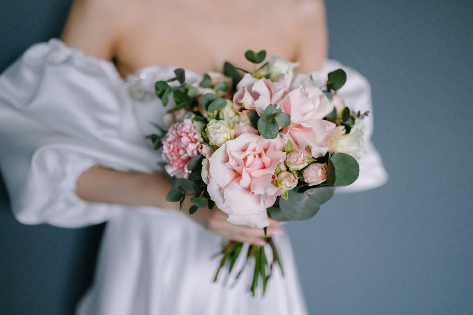 A person dressed in a white satin gown is holding a floral bouquet consisting of soft pink and white roses, pale pink peonies, and white lisianthus, accented with eucalyptus foliage. The bouquet is ar