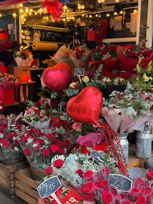 A vibrant floral display at a flower shop showcasing a variety of fresh red roses, white flowers, and greenery arranged in bouquets and wrapped in pink and white floral paper. Prominent in the arrange