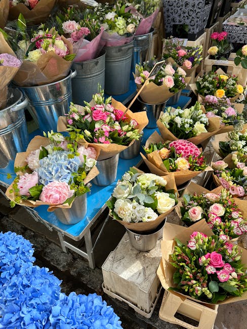 A display of fresh floral arrangements and bouquets at a flower market, featuring a variety of pastel and vibrant colors including pink roses, white hydrangeas, and light blue flowers, all wrapped in 