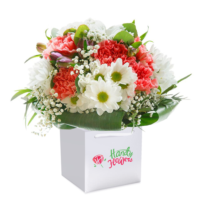 Upright in a sleek, square white gift bag, this expressive floral bouquet epitomises the style and vibrancy of Wood Green. Waves of open coral, peach, and red carnations form the heart of the arrangement, their soft, ruffled layers hinting at tactile richness and creative exuberance. Eye-catching white daisies brighten the bouquet with sunny lime-green centres, echoing the optimism found in local parks or bustling market stalls. Wisps of baby's breath bring an airy delicacy, weaving a gentle web through the bouquet's core. Amethyst-tinged unopened buds, poised on the left, inject subtle mystery and keep the eye moving. Underpinning the arrangement is an abundant collar of lush, glossy green foliage-its sheen and density recalling the greenery along Lordship Lane-providing not just a visual frame but a sense of constructed elegance. The meticulously rounded presentation, illuminated by deft, even lighting, radiates comfort and joy, promising a refreshing surprise sure to uplift any room. The sophisticated white gift bag, enhanced by secure ribbon handles, suggests a bouquet made for easy giving or swift delivery from Wood Green's trusted florists. Set against an uncluttered white background, the entire scene draws attention to florist expertise and product quality, making this bouquet a graceful gesture for celebrations, heartfelt thank-yous, or daily moments of happiness in the local area.